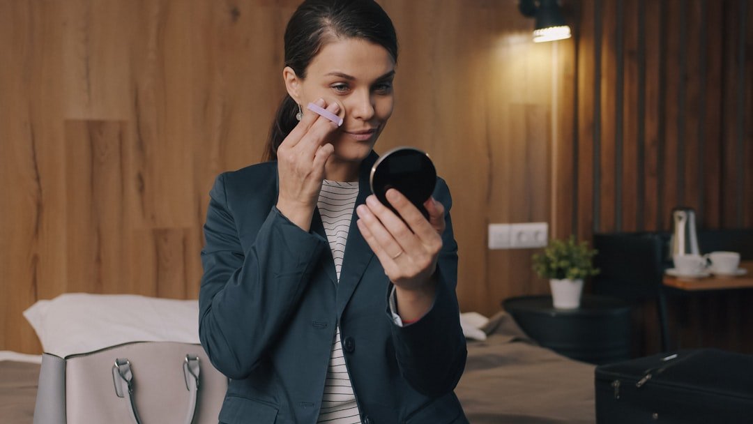 A woman in a blazer applies makeup to her cheek while looking into a compact mirror.