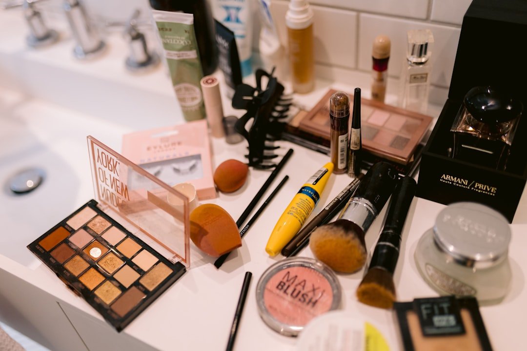 A bathroom counter covered with makeup products and tools, including eyeshadow palettes, brushes, mascara, blush, sponges, and fragrance.