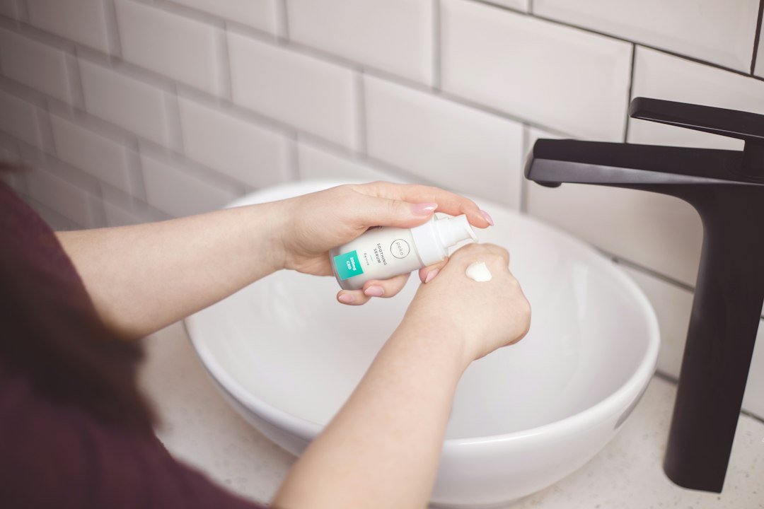 A person dispenses skincare cream from a pump bottle onto the back of their hand beside a bathroom sink.