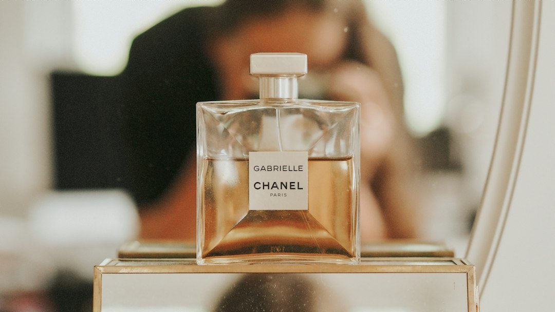 A clear glass bottle of Chanel Gabrielle perfume displayed on a mirrored vanity, with a blurred person in the background.