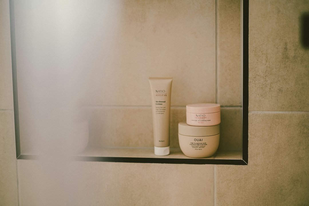 A skincare exfoliator, sleeping mask jar, and hair treatment jar arranged on a tiled bathroom shelf.
