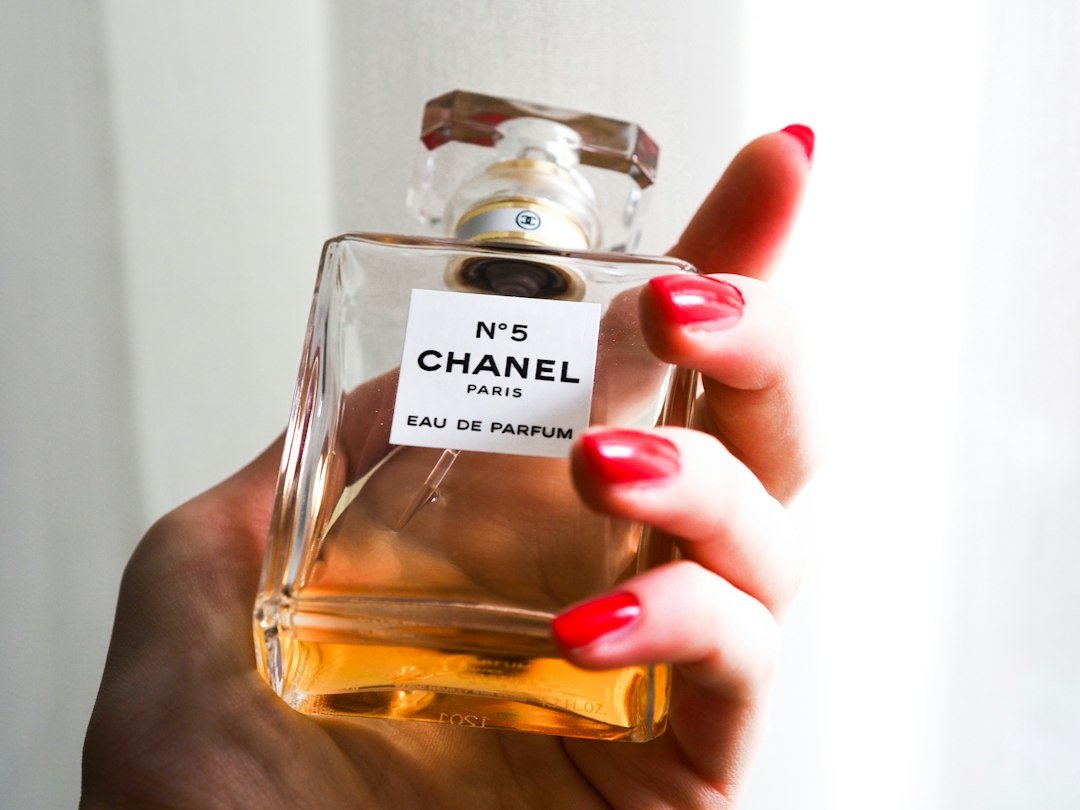 A hand with red-painted nails holds a clear bottle of Chanel N°5 eau de parfum.