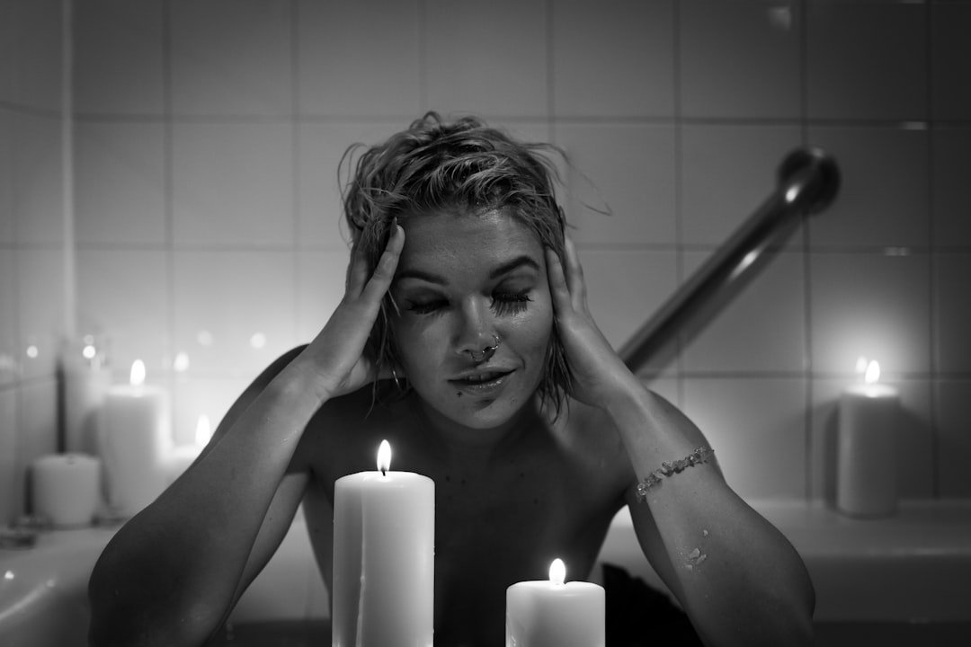 A person sits in a bathtub surrounded by lit candles, with hands resting against their head.