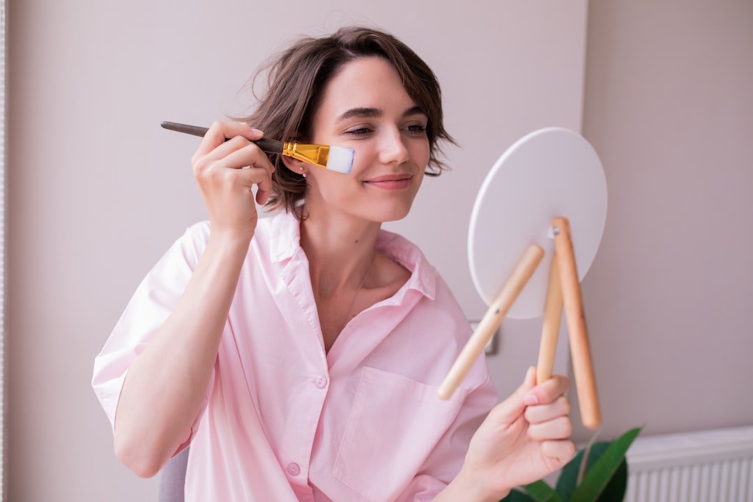 A woman applies complexion makeup to her cheek with a brush while looking into a tabletop mirror.