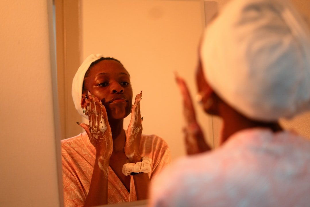 A person wearing a towel headband washes their face with foaming cleanser while looking in a bathroom mirror.