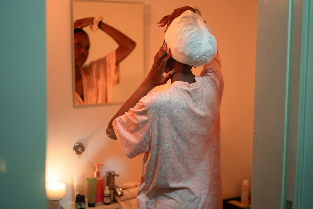 A person in a bathrobe and towel wrap touching their face and neck beside a bathroom sink with skincare products.