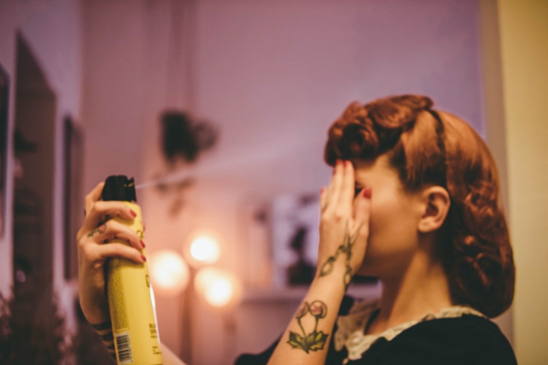 A person with styled, curled hair holds an aerosol hair product while shielding their face in front of a mirror.