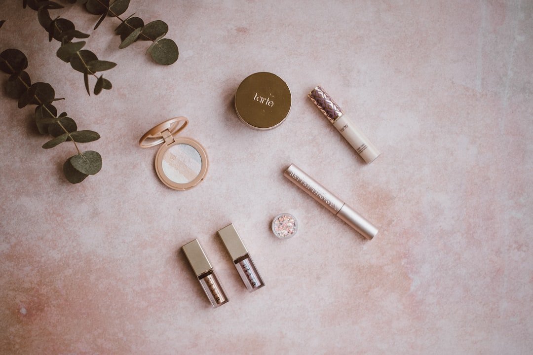 A curated flat lay of several makeup products, including compacts, tubes, and liquid cosmetics, arranged on a pale pink surface with eucalyptus leaves.
